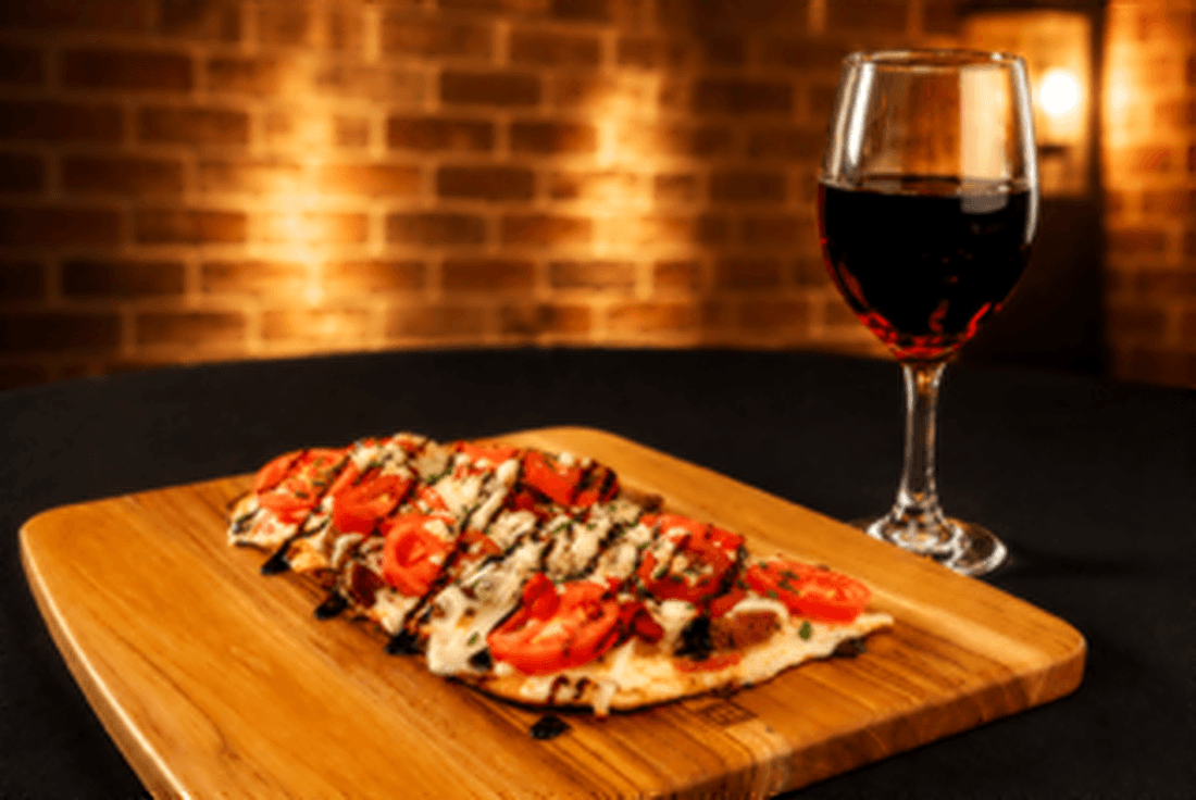 Stone-fired flatbread topped with tomato, cheese and balsamic glaze on a wood board beside a glass of red wine